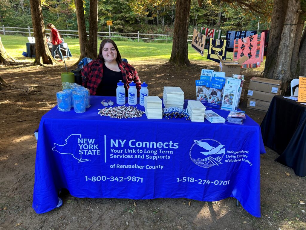 NY Connects staff, Erica Warnock, sits at a table, covered with blue NY Connects tablecloth covered with program information and branded items.